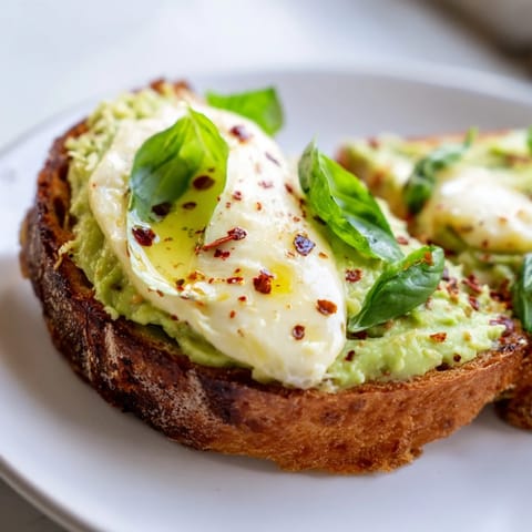 A close-up view of a warm avocado mozzarella tartine, featuring bubbly cheese and fresh basil garnish on crisp, golden-brown bread.